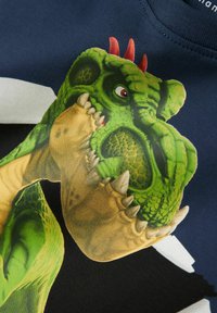 T-shirt blu navy con una grafica dettagliata di un dinosauro verde con spine rosse, artigli beige e un motivo di pelle testurizzata su uno sfondo scuro.