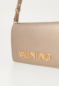 Borsa Valentino in pelle beige texturizzata con forma rettangolare. Presenta un logo dorato sul frontale e una tracolla dettagliata.
