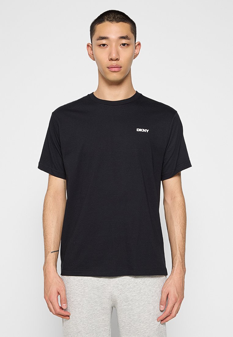 Mężczyzna ubrany w czarną koszulkę DKNY i jasnoszare spodnie stoi na tle jednolitego jasnego koloru.