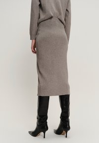 Persoon in een beige geribbelde midi-rok en bijpassende trui, met zwarte puntige hielknieboten, gezien vanuit de rug.