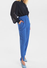 Kobieta ubrana w niebieskie spodnie o wysokim stanie z teksturą, z przednimi kieszeniami i rozcięciami na dole, w połączeniu z przezroczystą czarną bluzką w niebieskie kropki oraz niebieskimi szpilkami.