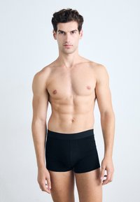 Zwarte katoen boxer shorts met een strakke pasvorm, brede elastische tailleband en stiksel details, model dat staat tegen een lichte achtergrond.