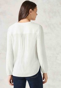 Blouse blanche à manches longues avec un tissu texturé à plis verticaux, un col rond et un ourlet arrondi. Portée avec un jean foncé.
