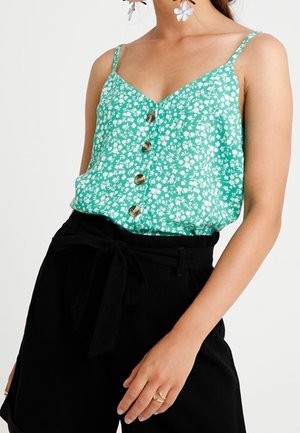 Woman wearing a green floral sleeveless top with buttons, black high-waisted pants tied with a belt, and flower-shaped earrings.