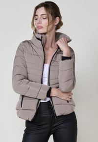 Pildīta bēša jakas ar augstu apkakli, ar rāvējslēdzēju aizdari un elastīgām piedurknēm. Mīksta oderes ar quilted (kviltētu) tekstūru un sānu kabatām.