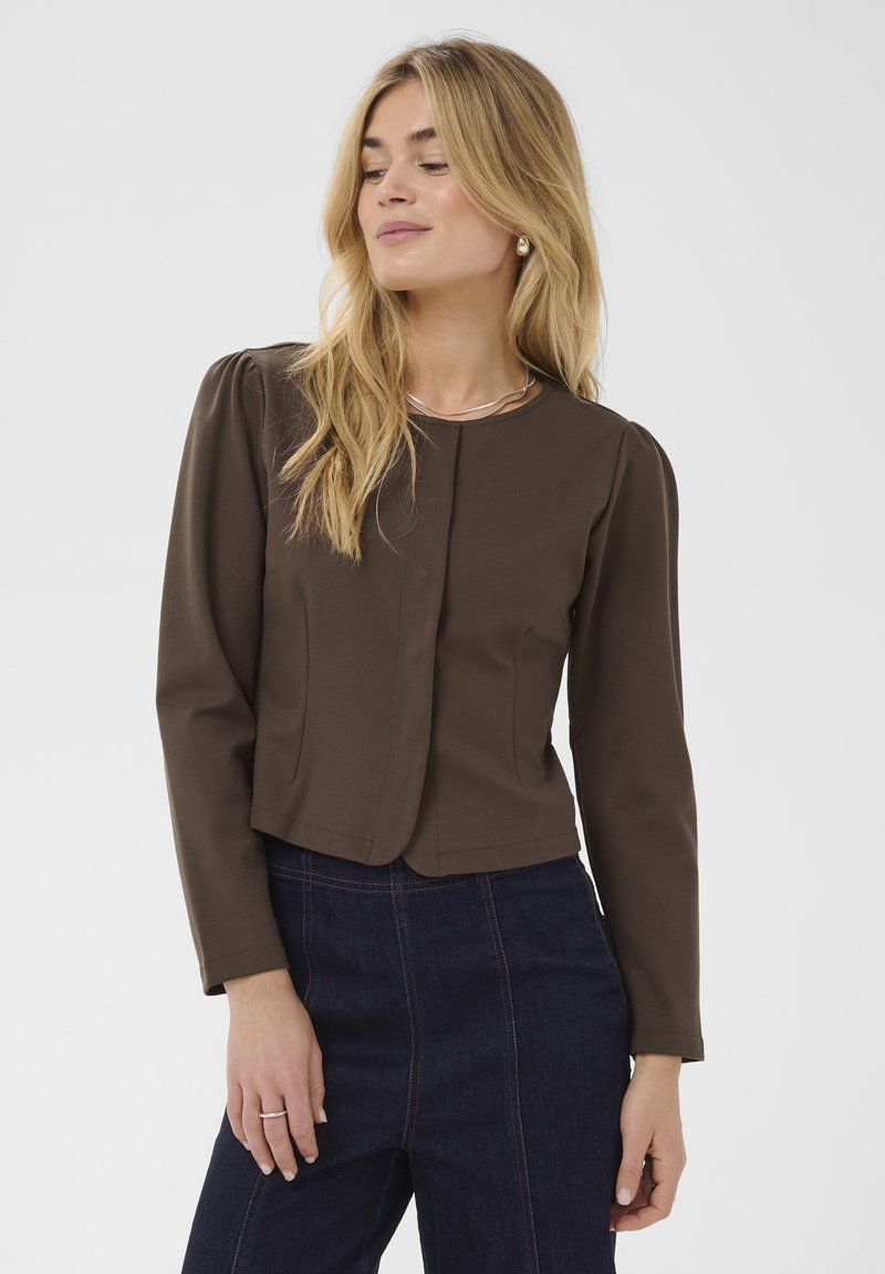 Femme blonde portant un chemisier marron à manches longues avec des épaules bouffantes et un jean taille haute bleu foncé sur un fond uni.