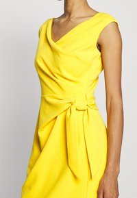 Gul klänning med V-ringning, korta kappärmar och knytning i midjan. Tillverkad av mjukt tyg med en slät textur och draperad design.