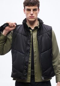 Νέος άνδρας με μαύρο γιλέκο καπιτονέ πάνω από πουκάμισο λαδί κρατά τον ιμάντα μιας τσάντας στον ώμο του, κοιτάζοντας ευθεία μπροστά με σοβαρή έκφραση.