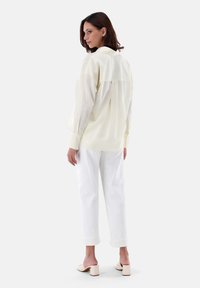 Blusa gialla chiara, strutturata, con colletto a camicia e design con parte posteriore arricciata, abbinata a pantaloni bianchi arrotolati e mule beige chiaro.