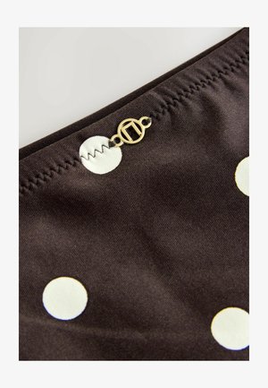 Pruun kangas valgete täpikestega, millel on volditud õmblus ja kuldne ümmargune sulgemisdetail. Suumvaade tekstiuri struktuurile.