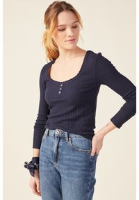 Maglietta a maniche lunghe rigata blu navy con scollatura ondulata e pattina con tre bottoni. Abbinata a jeans azzurri a vita alta.