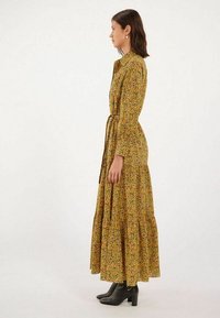 Robe longue à fleurs avec un fond jaune, présentant un design à volants, des manches longues et une ceinture à la taille. Associée à des bottines noires.