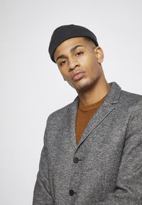 Blazer gris texturé avec des boutons noirs sur un pull en maille orange, complété par un bonnet noir. Design moderne et ajusté.