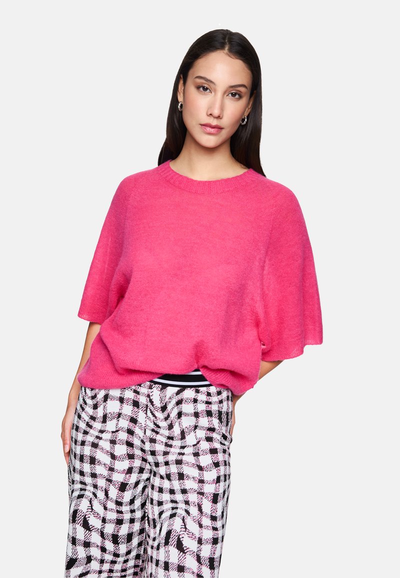 Rosa oversized Strickpullover mit Rundhalsausschnitt, kurzen Ärmeln und weicher Textur; kombiniert mit schwarz-weißen karierten Hosen.