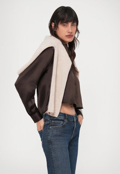 Sötét hajú nő barna, rövid szatén ingben, kék farmerben és vállára terített világos pulóverrel, kezét a zsebében tartva.