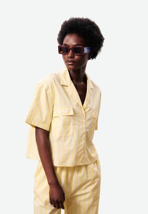 Vrouw met kort krullend haar, die oversized rechthoekige zonnebrillen draagt en een loszittende gele gestreepte korte mouwen blouse en bijpassende broek.