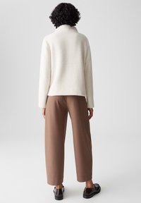 Maglione bianco testurizzato con colletto alto, abbinato a pantaloni marroni a gamba larga e scarpe nere lucide. Design minimalista e tonalità neutre.