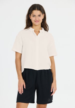 Kortärmad, lätt krämfärgad blus med krage, i kombination med svarta, mönstrade shorts. Har en något beskuren design och en avslappnad passform.