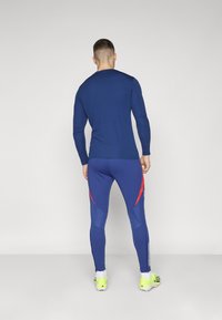 Marinblå långärmad topp med rund halsringning; figurnära marinblå leggings med röda accenter och vita ränder på sidorna; ljusgula sneakers.
