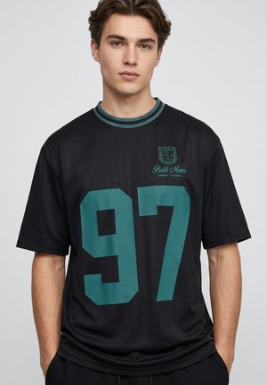 Young man wearing a black short-sleeve shirt with green "97" and "Bold Move Urban Studio" text, standing with hands in pockets.