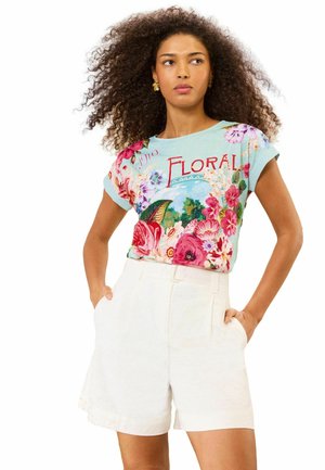 Femme aux cheveux bouclés portant une chemise à manches courtes à motif floral avec le texte « FLORAL » et un short blanc taille haute, mains dans les poches.