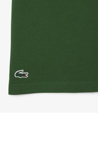 Camiseta de algodón verde con una textura suave, que presenta un pequeño logotipo bordado de cocodrilo en blanco y rojo en el bajo izquierdo.