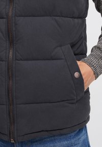 Chaleco acolchado navy con cremallera frontal, que cuenta con un bolsillo lateral y un botón a presión en bronce, combinado con un suéter de punto texturizado de manga larga en gris.
