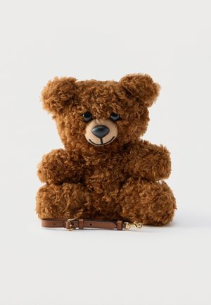 Orso di peluche marrone riccio seduto con occhi e naso neri, sorridente, con una cinghia di pelle marrone staccata davanti su sfondo bianco.