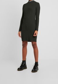 Robe côtelée vert foncé à manches longues et col montant, avec un accent rayé blanc au niveau du col, associée à des bottes de combat noires.