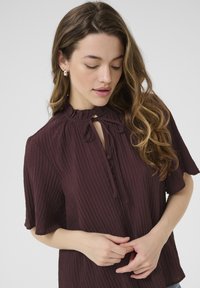 Bordeaux blouse met een lichtgewicht, geribbelde textuur. Kenmerkt zich door een rimpelrand bij de hals, een strikdetail en knopen aan de voorkant. Korte mouwen en een relaxte pasvorm.