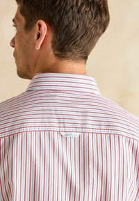 Košela s golierom s vertikálnymi červenými a bielymi pruhmi, s malou etiketou s nápisom "JOULES" na zadnej časti výstrihu. Bavlnený materiál, hladká textúra.