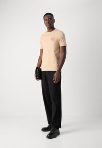 Helles Pfirsich-T-Shirt mit roten grafischen Akzenten, kombiniert mit schwarzen Hosen und schwarzen Schuhen, gehalten mit einer schwarzen Tasche. Glatte Stofftextur.