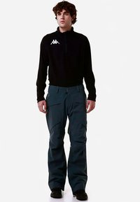 Top a maniche lunghe nero con zip e logo bianco, abbinato a pantaloni da sci verde petrolio scuro con tasche cargo e vestibilità comoda. Scarpe nere.