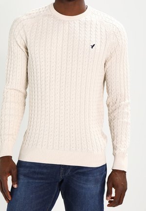 Homme portant un pull crème en maille torsadée avec poignets et ourlet côtelés, associé à un jean bleu foncé.