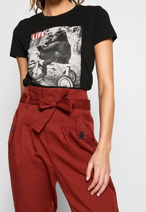 Person wearing a black t-shirt with a gorilla on a tricycle and the word LIFE, paired with high-waisted rust-red pants tied with a bow belt.