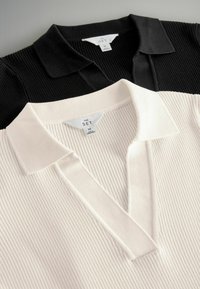 Twee poloshirts met kraag tentoongesteld: de ene in zwart met ribbeltextuur, de andere in crème, met een vergelijkbaar ribbelpatroon en labels.