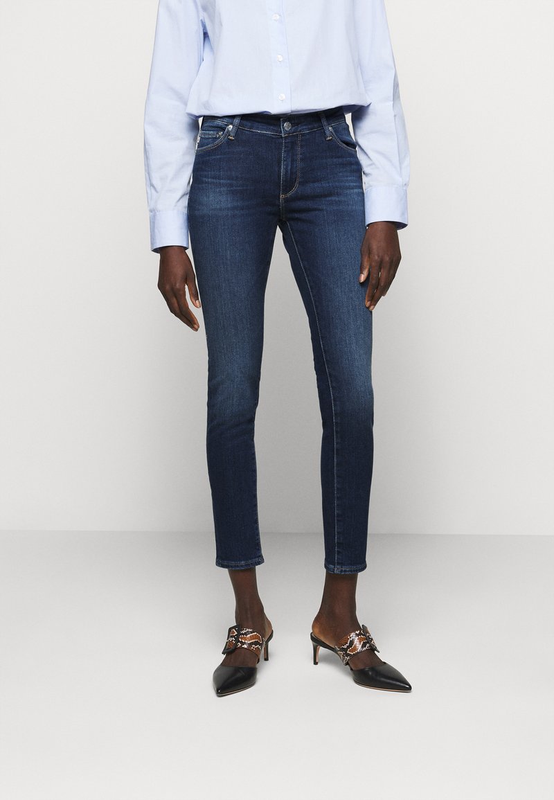 Jeans skinny azul escuro com um leve desbotamento, combinados com uma camisa azul clara de botões e sapatos de salto agudo pretos com detalhes em castanho e bege.