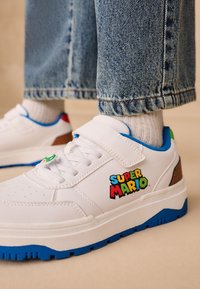 Baskets blanches avec semelles bleues et une bride à Velcro, arborant un logo coloré "Super Mario" et des accents marron au talon.