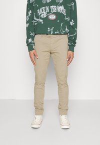 Chinos beige avec une coupe ajustée, en coton. Portés avec un sweatshirt vert arborant un graphique blanc et du texte. Des baskets blanches complètent la tenue.