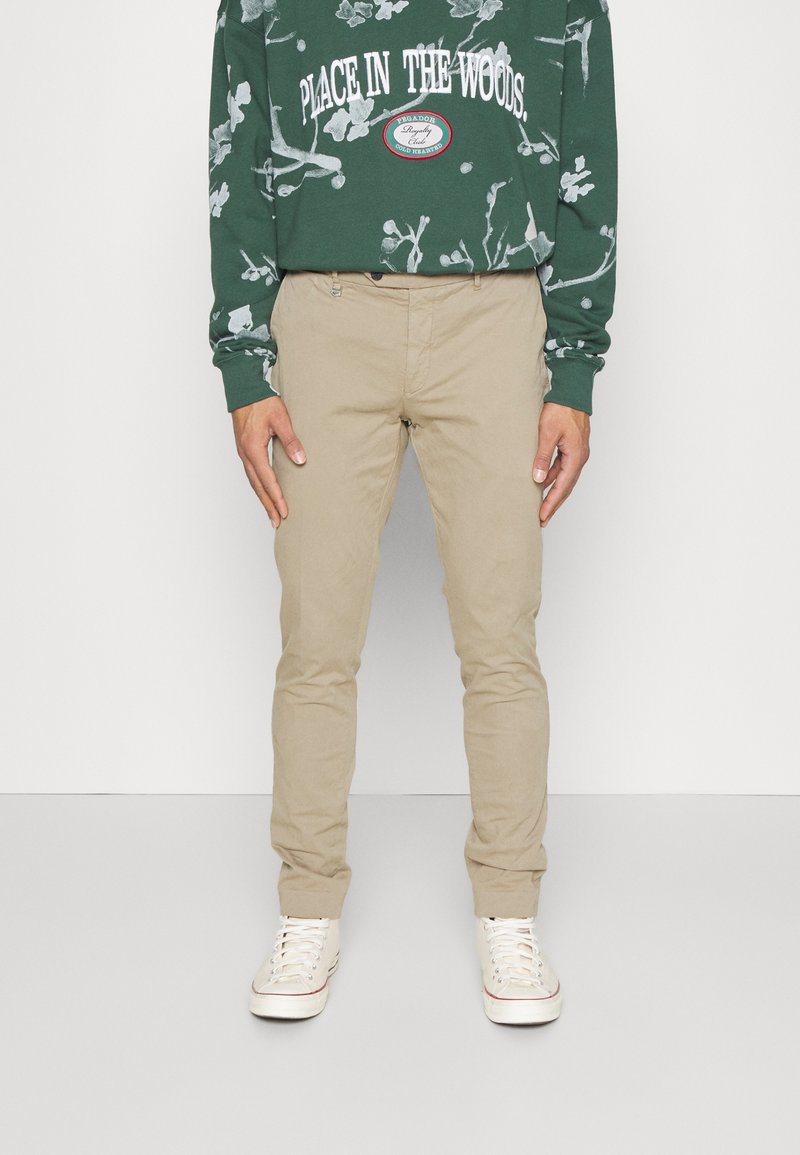 Chinos beige avec une coupe ajustée, en coton. Portés avec un sweatshirt vert arborant un graphique blanc et du texte. Des baskets blanches complètent la tenue.
