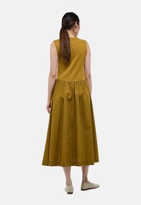 Femme avec une longue queue de cheval brune portant une robe midi sans manches de couleur moutarde et des ballerines beiges, se tenant dos à l'appareil photo sur fond blanc.