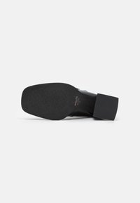 Chaussure en cuir noir avec bout carré et talon bloc, semelle présentant une surface antidérapante et le nom de la marque « Minelli » imprimé près du talon.