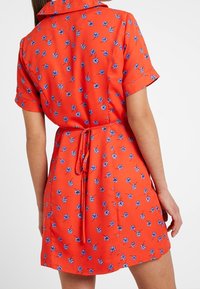 Röd blommig klänning med korta ärmar, prydd med blå blommönster, med en åtsittande midja och en knytning på baksidan. Mjuk, lättviktig tyg.