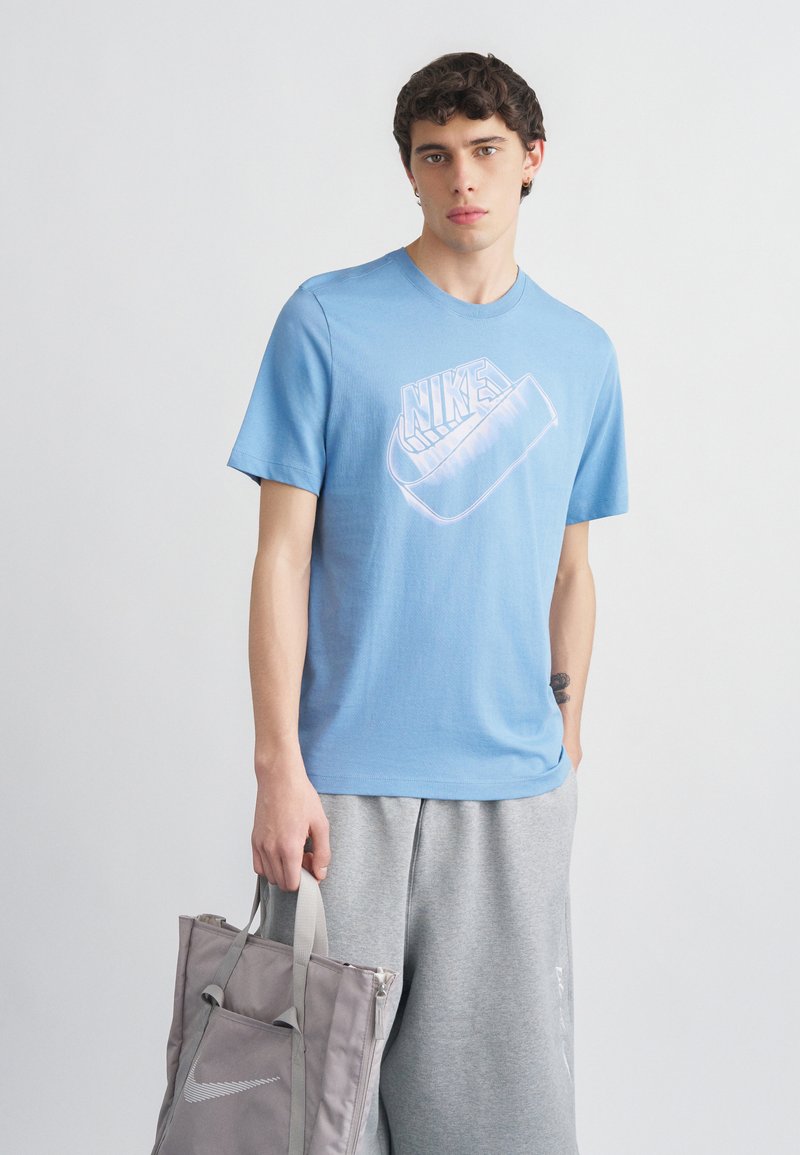 Jeune homme portant un t-shirt Nike bleu et un pantalon de survêtement gris, tenant un sac fourre-tout gris avec le logo Nike sur un fond uni.