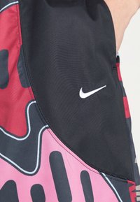 Nike athletic shorts in black, pink, and gray features a wavy pattern, white logo, and smooth, textured fabric. Accents enhance design.