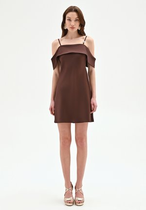 Femme portant une robe chocolat marron sans manches, décolletée avec bretelles spaghetti et sandales à talons hauts métalliques dorées, debout droite.