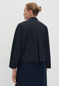 Navy cropped jacket with a pleated back, long sleeves, and a standing collar. Made from smooth, textured fabric.