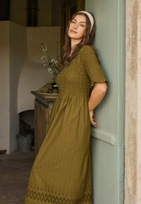 Ženska z dolgimi rjavimi lasmi, oblečena v zeleno teksturirano midi obleko in belo trak za lase, se naslanja ob bledo zeleno vrata v rustikalni sobi.