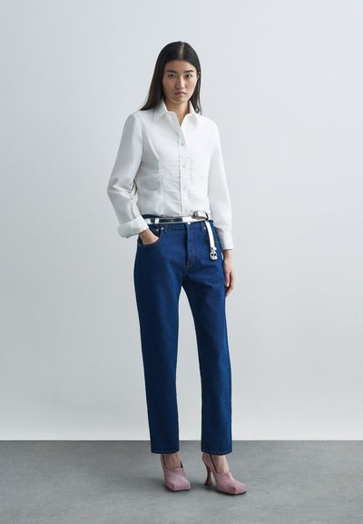 Λευκό πουκάμισο με μακριά μανίκια και λεπτομέρειες συγκέντρωσης μπροστά, συνδυασμένο με μπλε τζιν παντελόνι ίσιας γραμμής. Το μοντέλο φοράει ροζ ψηλοτάκουνες γόβες.