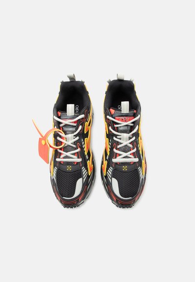 Ζευγάρι αθλητικών παπουτσιών σε μαύρο, κόκκινο και κίτρινο χρώμα με λευκά κορδόνια και πορτοκαλί πλαστική ταμπέλα στο αριστερό παπούτσι, φωτογραφημένα από πάνω σε λευκό φόντο.